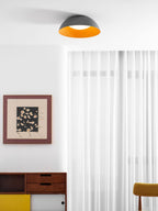 Lit grey and orange Clérachic ceiling light above a white room with long sheer curtains, a framed print and a wooden sideboard.