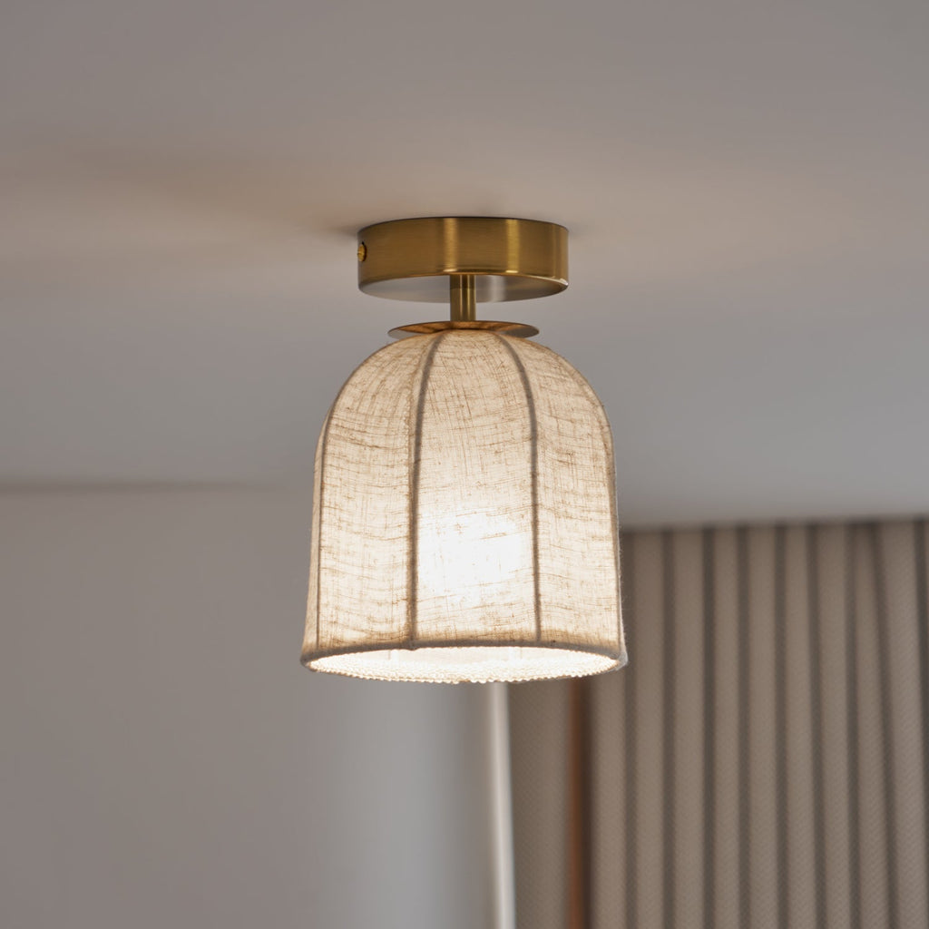 A warmly lit Clérachic linen-shade ceiling light with a gold canopy, shown near a curtain and a pale grey ceiling.