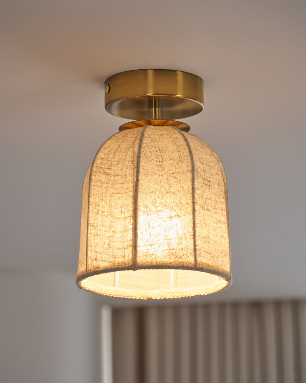 A Clérachic ceiling light with a warm lit linen shade and a gold canopy, seen at an angle with soft curtains behind.