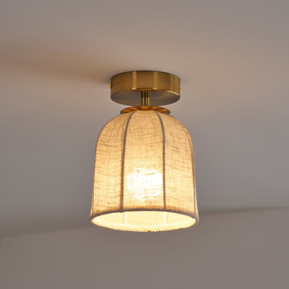 Smaller Clérachic ceiling light glowing warm, with its linen shade and gold canopy set against a plain grey ceiling.