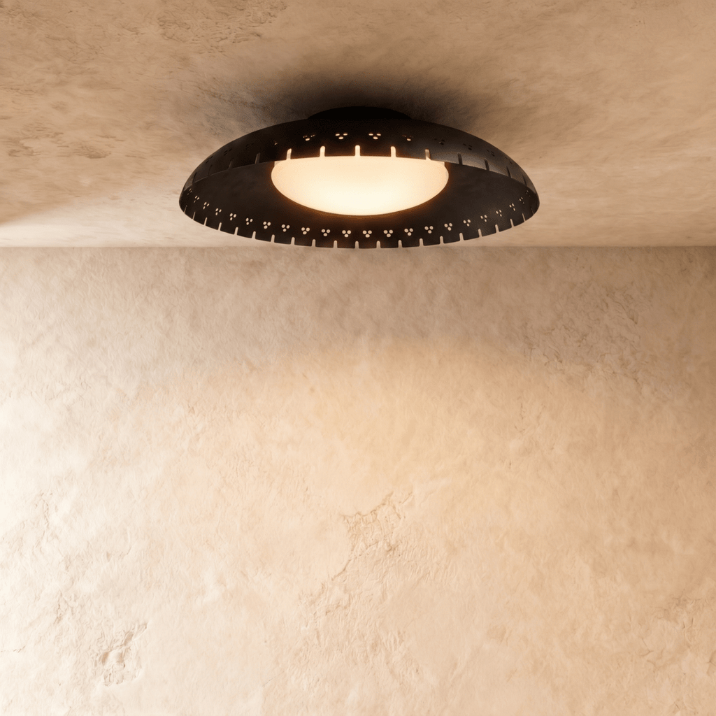 Clérachic ceiling light in matte black, a shallow perforated drum glowing warmly against a plaster ceiling.
