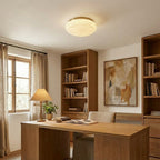 A gold-rimmed Clérachic ceiling light hangs low over a wooden desk, with a table lamp and bookshelves beside it.
