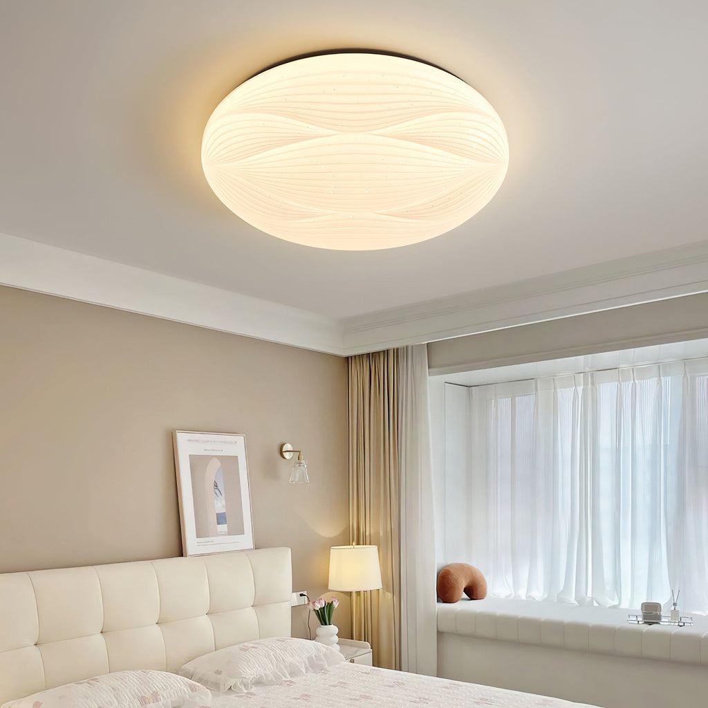 A wide, lit Clérachic ceiling light with a black rim above a tufted cream bed and a window seat.