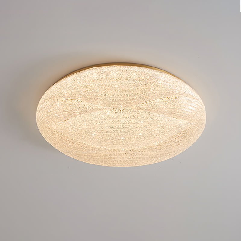 A wider gold-rimmed Clérachic ceiling light lit with a glittering textured shade against a grey ceiling.
