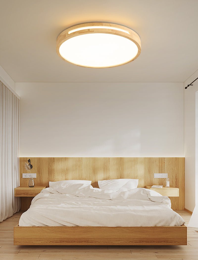 Round ceiling light from Clérachic with a light wood frame, lit, above a low wooden bed with white linen in a plain bedroom.