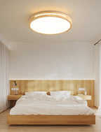 Round ceiling light from Clérachic with a light wood frame, lit, above a low wooden bed with white linen in a plain bedroom.