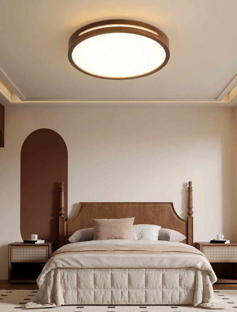 A round walnut-frame Clérachic ceiling light lit above a carved headboard bed, with rattan bedside tables on each side.