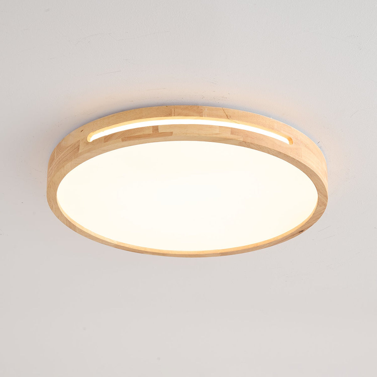 Round pale wood ceiling light from Clérachic, lit warm, with a glowing slot above the frosted panel on a grey ceiling.