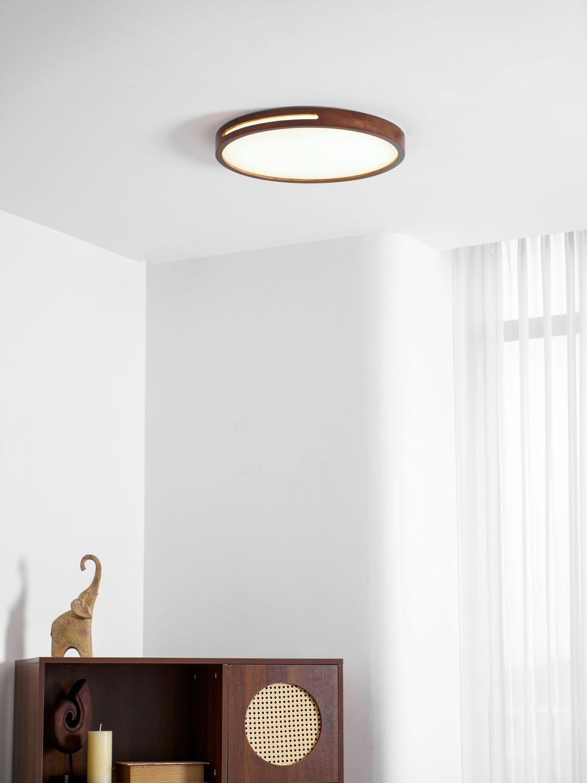 A small round walnut ceiling light from Clérachic in a white room with sheer curtains and a dark wooden cabinet.