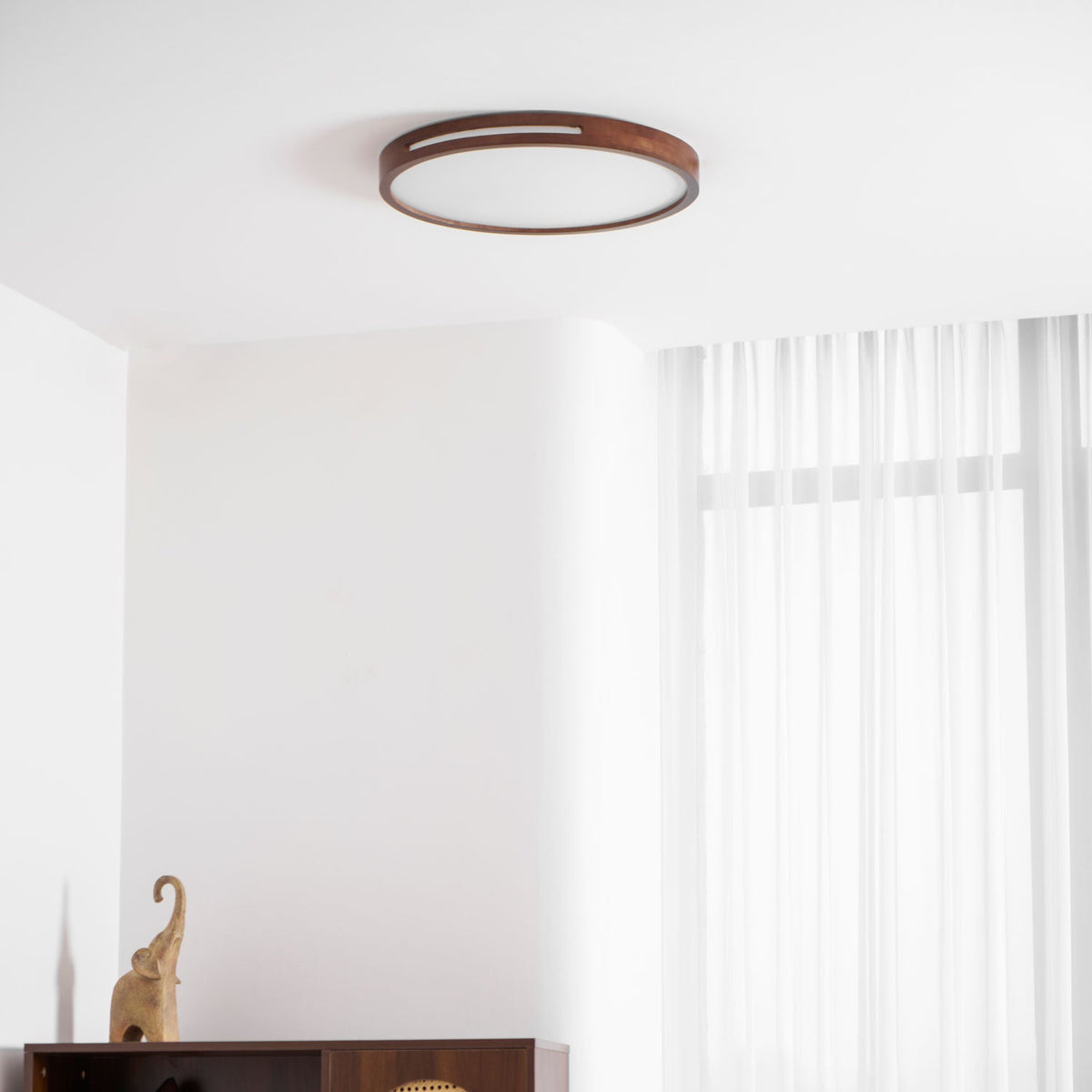 Round walnut-frame ceiling light from Clérachic seen high on a white ceiling beside sheer curtains and a carved figurine.
