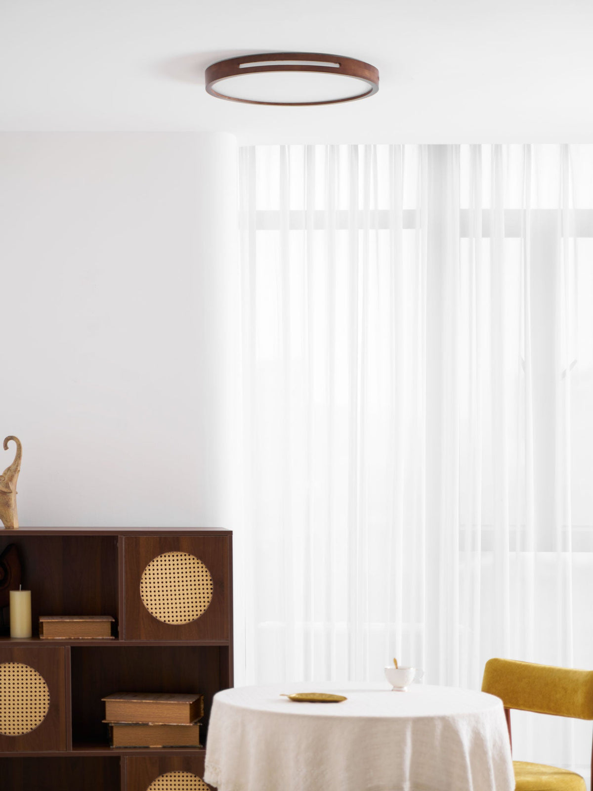Round Clérachic ceiling light in walnut, lit, above a corner with a rattan-front shelf unit and a linen-covered table.