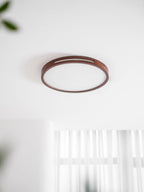 Walnut ring ceiling light from Clérachic on a white ceiling, with a blurred curtain and a leaf at the edge of the frame.