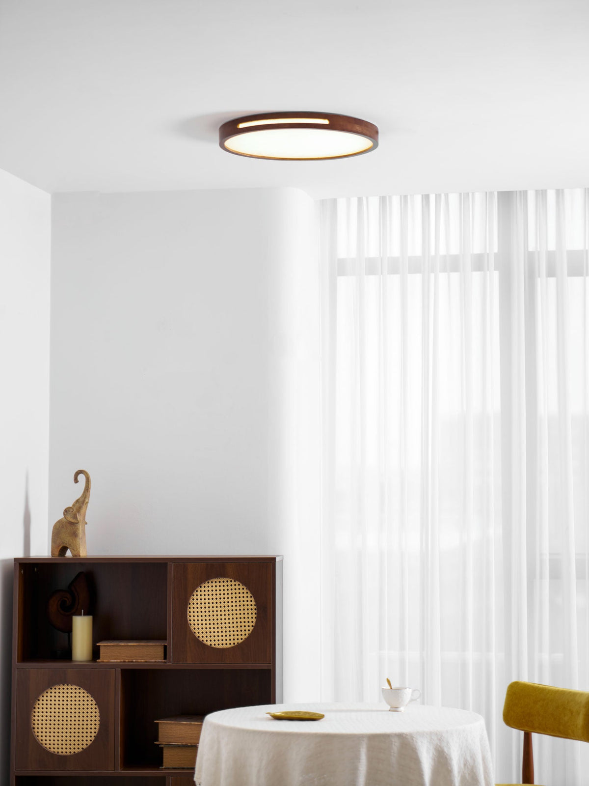 Round walnut ceiling light from Clérachic glowing softly over a dining corner with a yellow chair and a rattan cabinet.