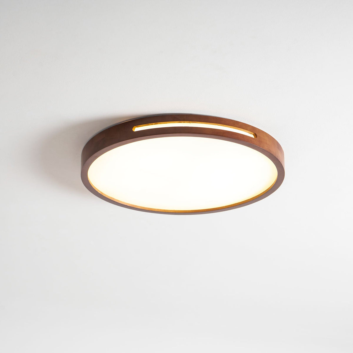 Side-on view of a lit round walnut Clérachic ceiling light, with a warm strip of light through the slot in its frame.