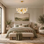The Clérachic petal chandelier glows above a bed with a sage throw and wooden bench, in a pale panelled bedroom.
