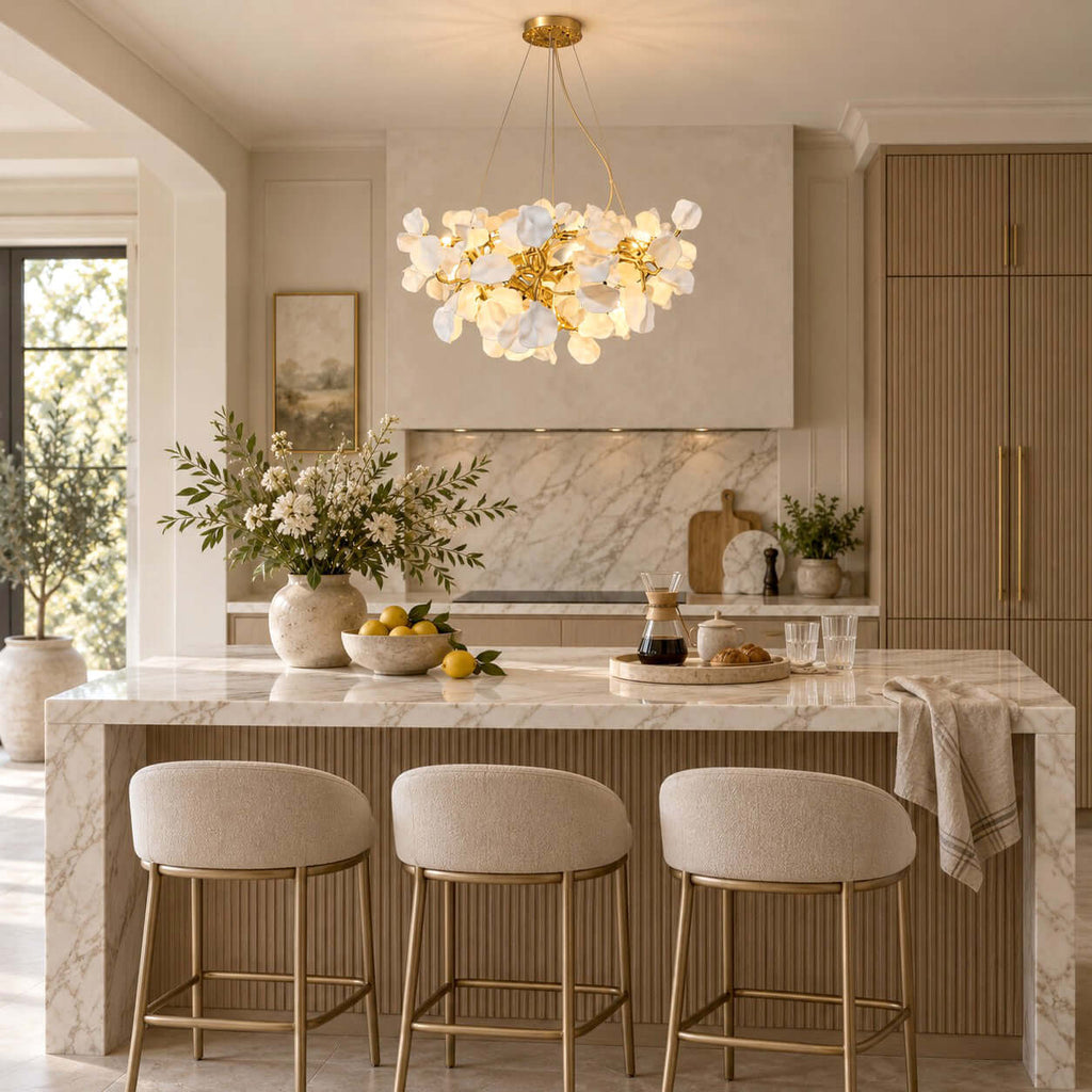 A lit Clérachic chandelier hangs over a kitchen island with a veined marble top and three upholstered brass-legged stools.