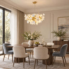 A Clérachic chandelier of white ceramic petals hangs above a wooden dining table with grey and cream chairs in a high-ceilinged room.