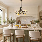 Lit Clérachic chandelier with ceramic blooms hung over a kitchen island with cream bar stools, a marble splashback behind.