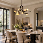 A Clérachic chandelier glowing above a marble dining table with upholstered chairs, tall glass doors and a landscape painting behind.