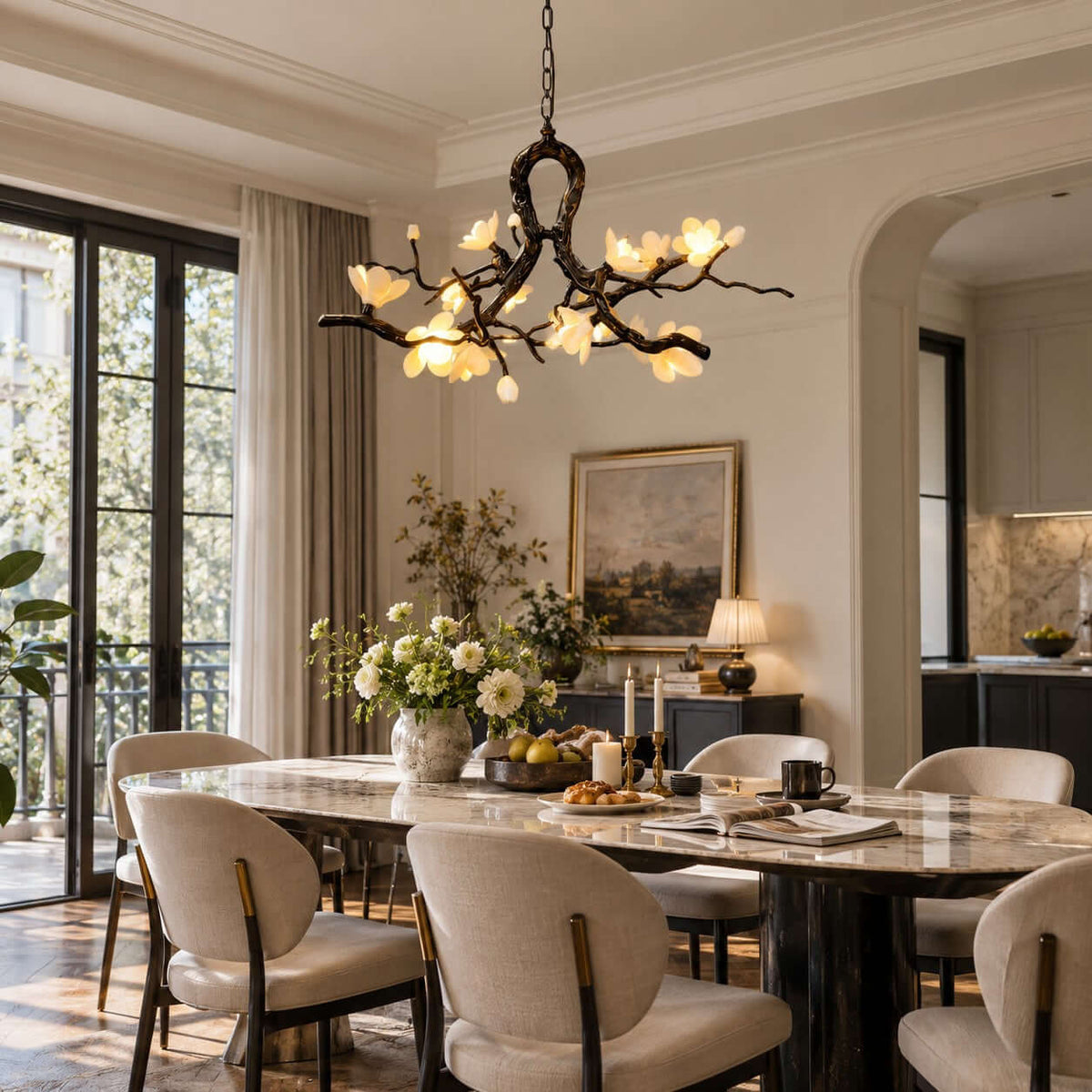 A Clérachic chandelier glowing above a marble dining table with upholstered chairs, tall glass doors and a landscape painting behind.