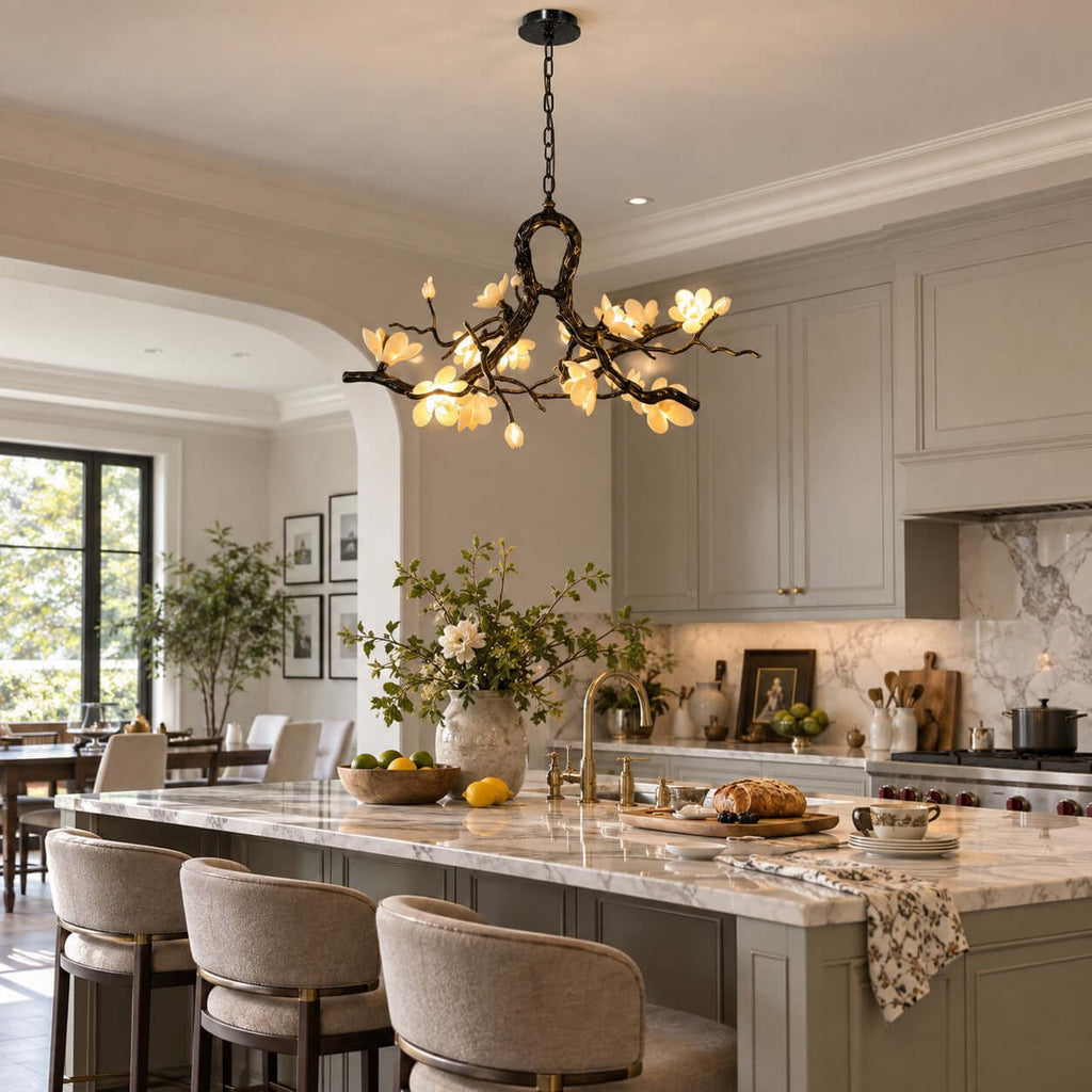 Clérachic chandelier lit above a long island with cream stools, grey-green cabinets and a marble splashback beyond.