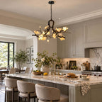 Clérachic chandelier lit above a long island with cream stools, grey-green cabinets and a marble splashback beyond.