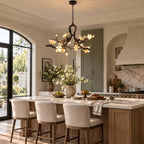 A Clérachic blossom chandelier above a kitchen island with wood-framed stools, in front of an arched window and a sage cupboard.