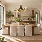 Clérachic chandelier lit above a plank dining table with six slipcovered chairs on a jute rug, a tall olive tree in the corner.