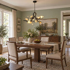 Lit Clérachic chandelier in a sage-green dining room, over a dark wood table with upholstered chairs and a landscape painting.