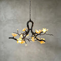 Lit Clérachic chandelier against a grey plaster wall, its dark vine frame glowing with warm yellow ceramic blooms and a few buds.