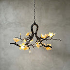 Lit Clérachic chandelier against a grey plaster wall, its dark vine frame glowing with warm yellow ceramic blooms and a few buds.