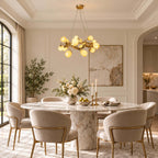 A Clérachic flower chandelier in a panelled cream dining room, glowing above a marble table with cream chairs trimmed in brass.