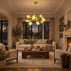 Lit at dusk above a dark wooden coffee table, this Clérachic gold chandelier glows in a living room with a fireplace and tall windows.