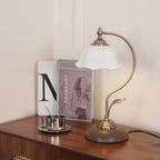 This Clérachic table lamp lit on a dark wood sideboard, beside a lookbook, a book with a large letter and a steel cup.