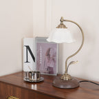Low view of this Clérachic table lamp on a wooden cabinet, its curved arm and round wood-grain base beside a stack of books and a cup.