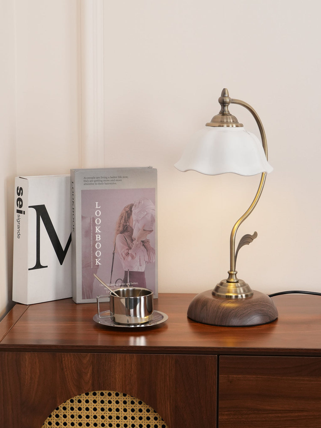 A Clérachic table lamp with a scalloped white shade on a wooden console with a rattan panel, glowing beside books and a small steel cup.
