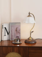 A Clérachic table lamp with a scalloped white shade on a wooden console with a rattan panel, glowing beside books and a small steel cup.