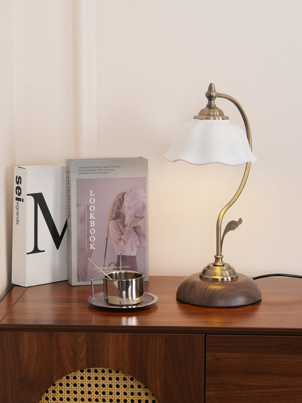 A Clérachic table lamp with a scalloped white shade on a wooden console with a rattan panel, glowing beside books and a small steel cup.