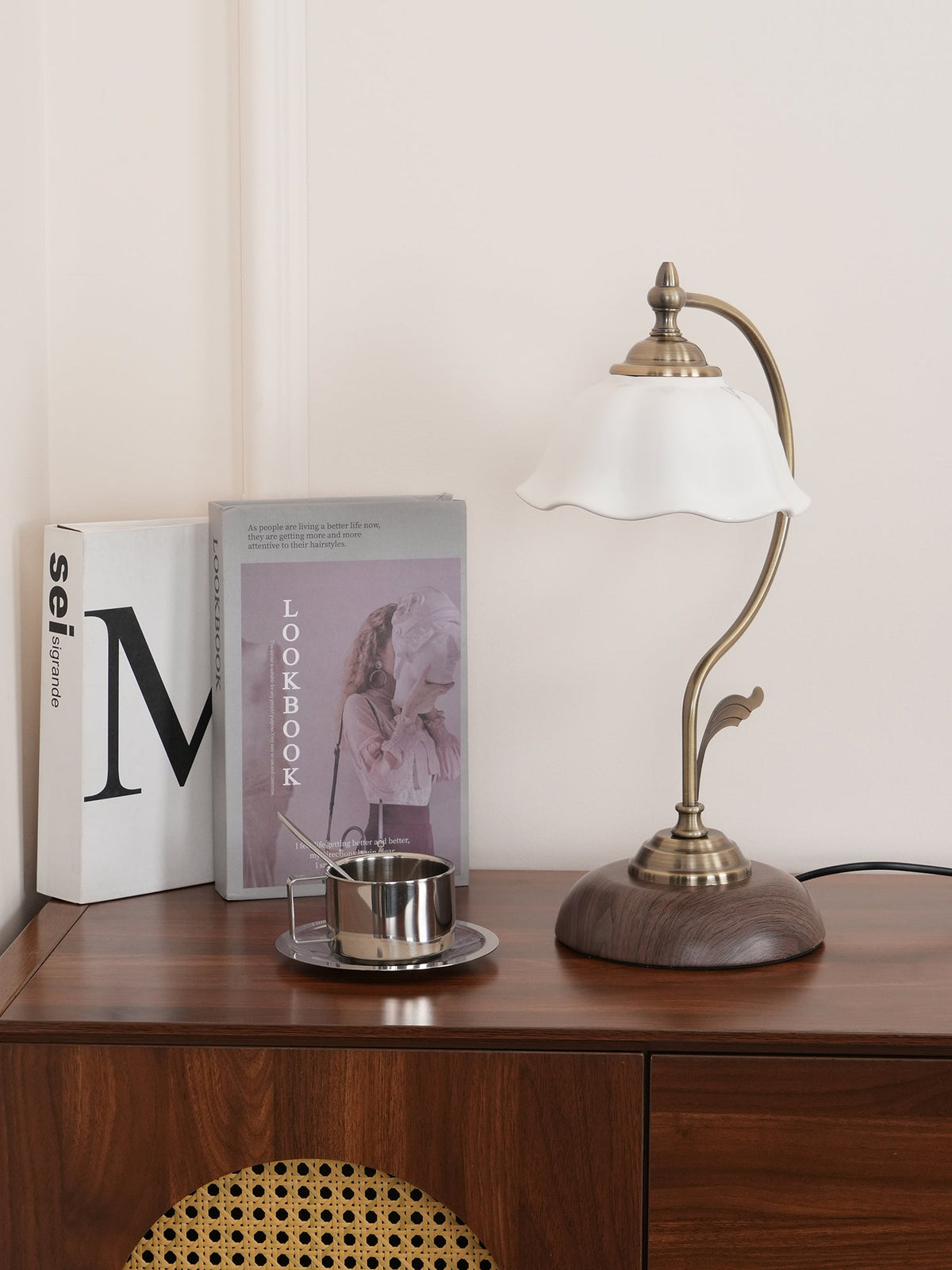 Clérachic table lamp unlit on a wood console, its white shade and bronze-toned arm in front of books on a cream wall.