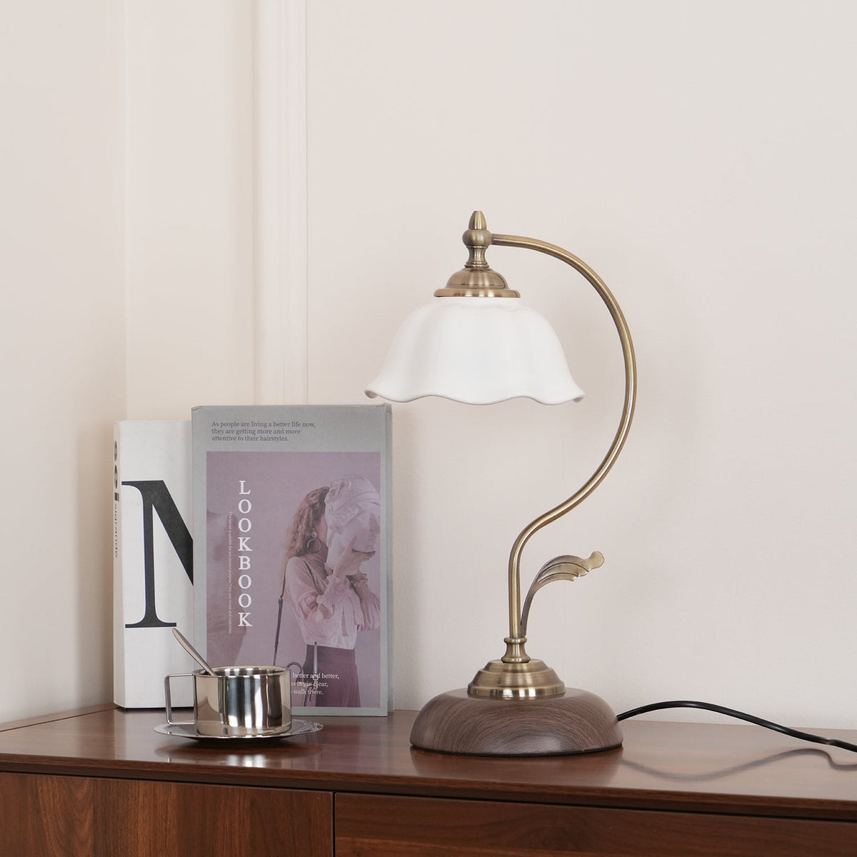 Clérachic lamp with a fluted white ceramic shade beside a lookbook and a steel cup on a dark wood sideboard.