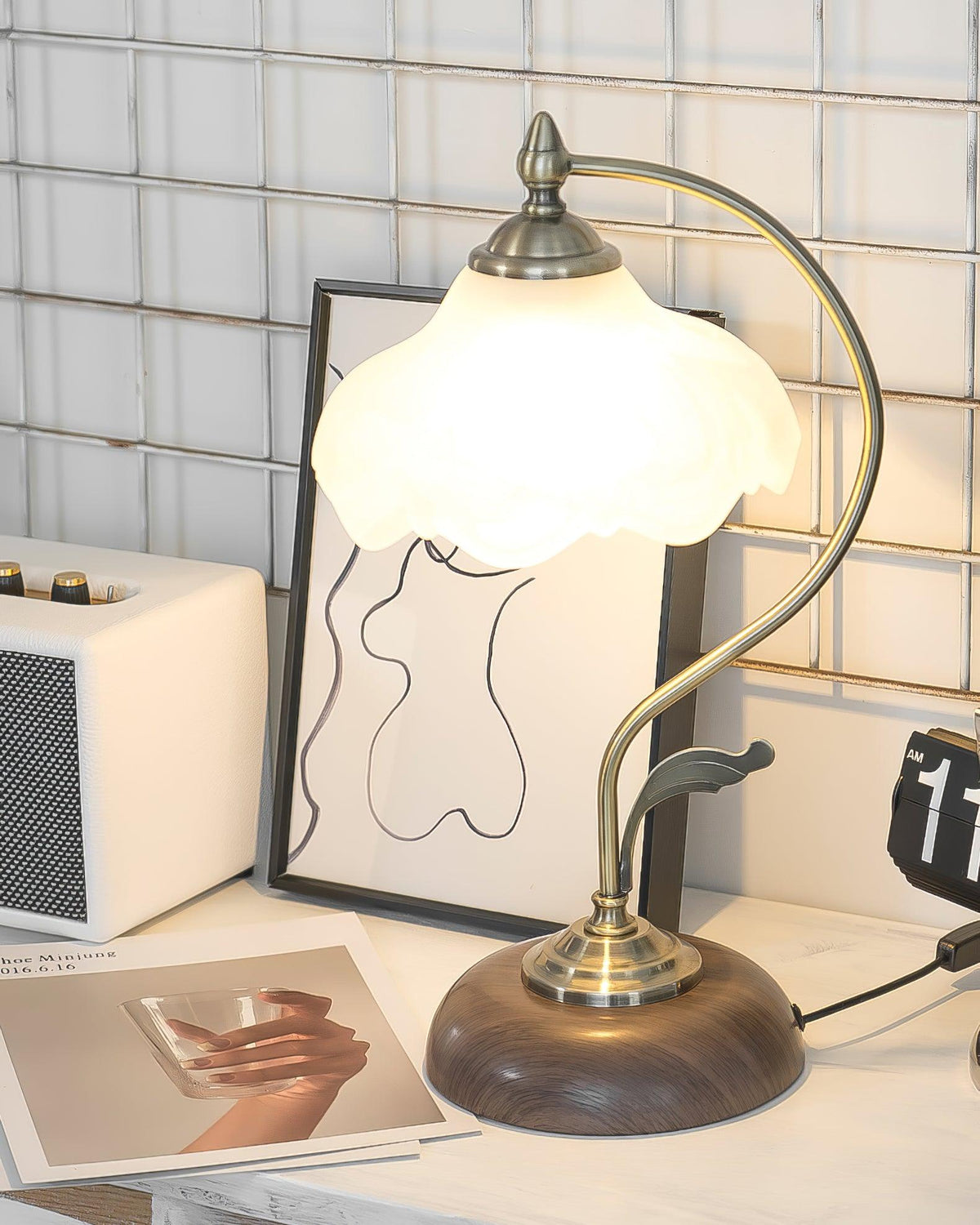 Lit Clérachic table lamp with a glowing frosted shade, set on a white desk in front of a framed line drawing and a cream speaker.