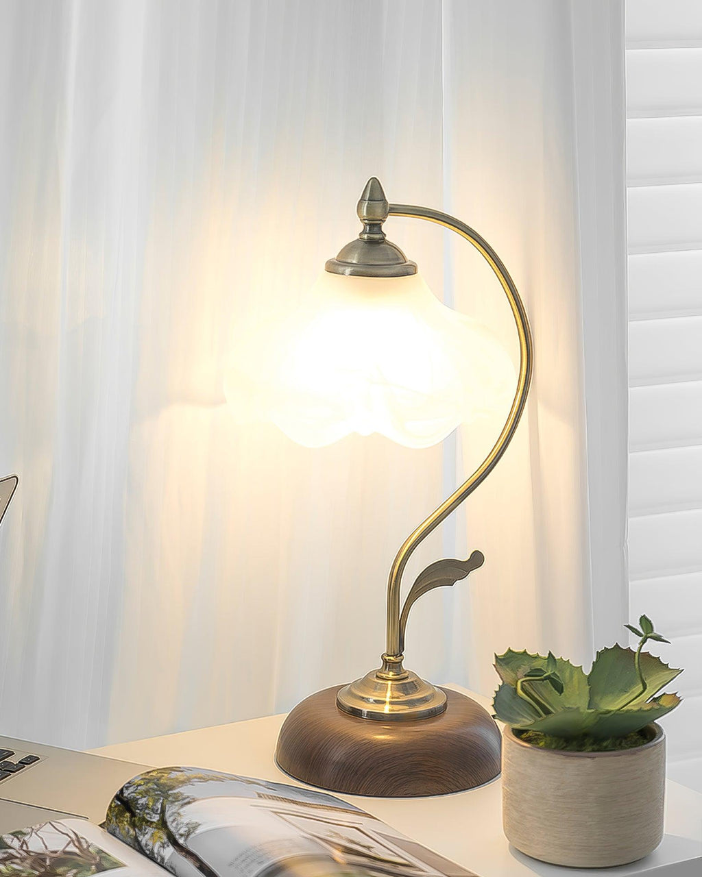 Frosted shade of this Clérachic table lamp aglow against a sheer curtain, with a succulent in a woven pot and a magazine below.