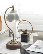 Unlit Clérachic table lamp next to a bronze bird on a black block and a ring ornament, on a shelf near a window.