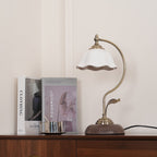 Clérachic table lamp with a white ceramic shade, standing on a wooden sideboard in front of a clothing book and a lookbook.