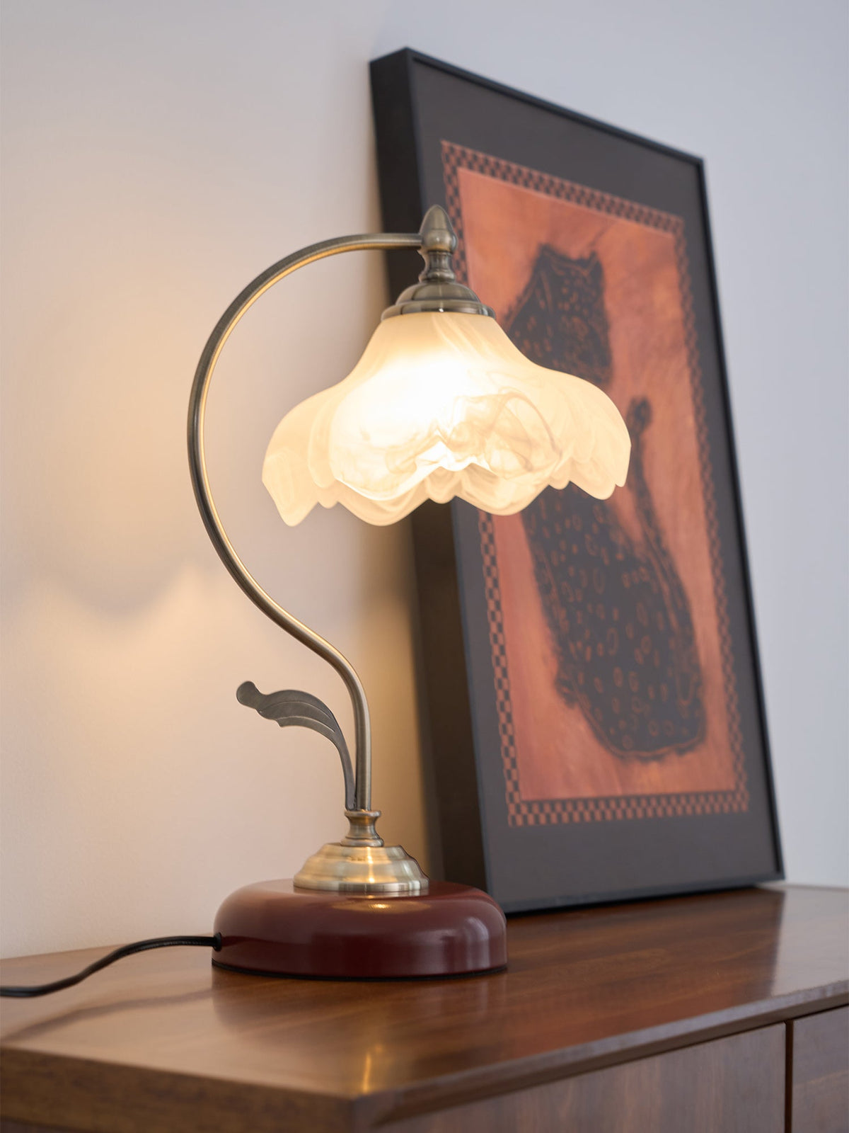 Lit Clérachic table lamp with a frosted glass shade, in front of a large framed print of a dark cat on an orange ground.