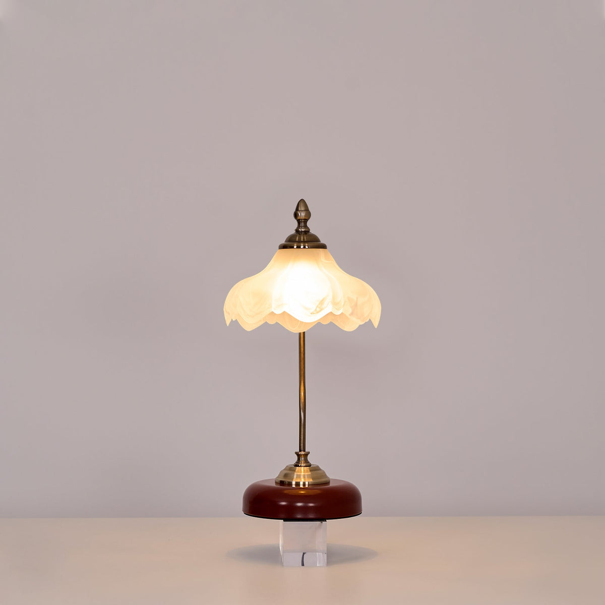 Lit Clérachic table lamp with a straight stem and a warm glowing glass shade on a clear block against a soft grey wall.