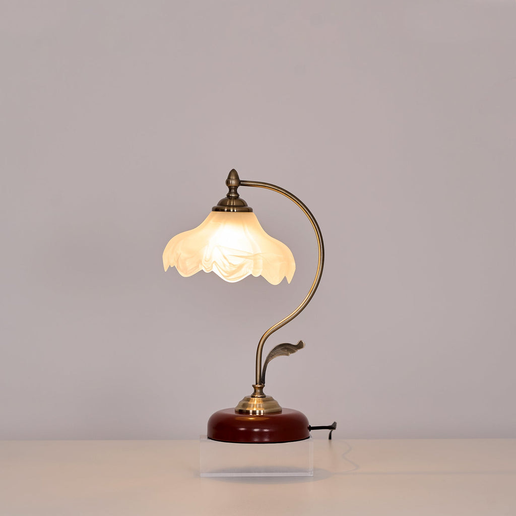 Clérachic table lamp lit against a grey wall, its curved arm and leaf detail rising from a round base on a clear acrylic plinth.