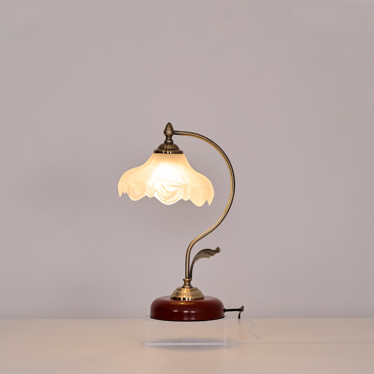 Clérachic table lamp lit against a grey wall, its curved arm and leaf detail rising from a round base on a clear acrylic plinth.