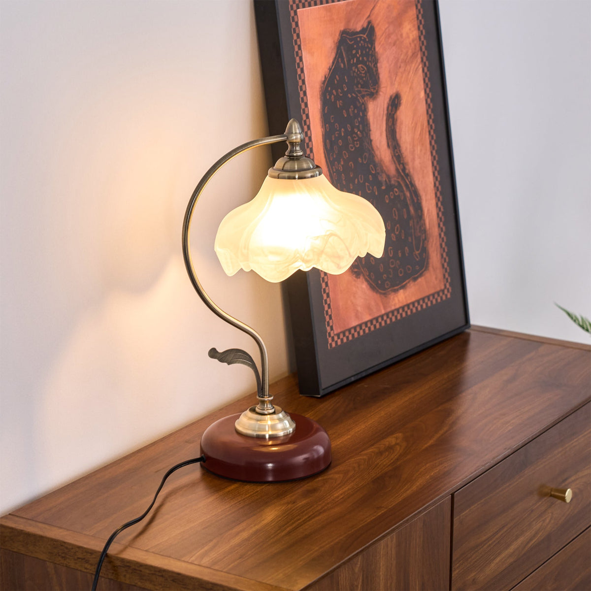 Lit Clérachic table lamp on a dark wood chest of drawers, beside a framed cat print and a cord trailing down the side.