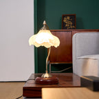 Clérachic table lamp with its frosted glass shade lit, on a wooden table in a room with a green wall and a grey armchair.